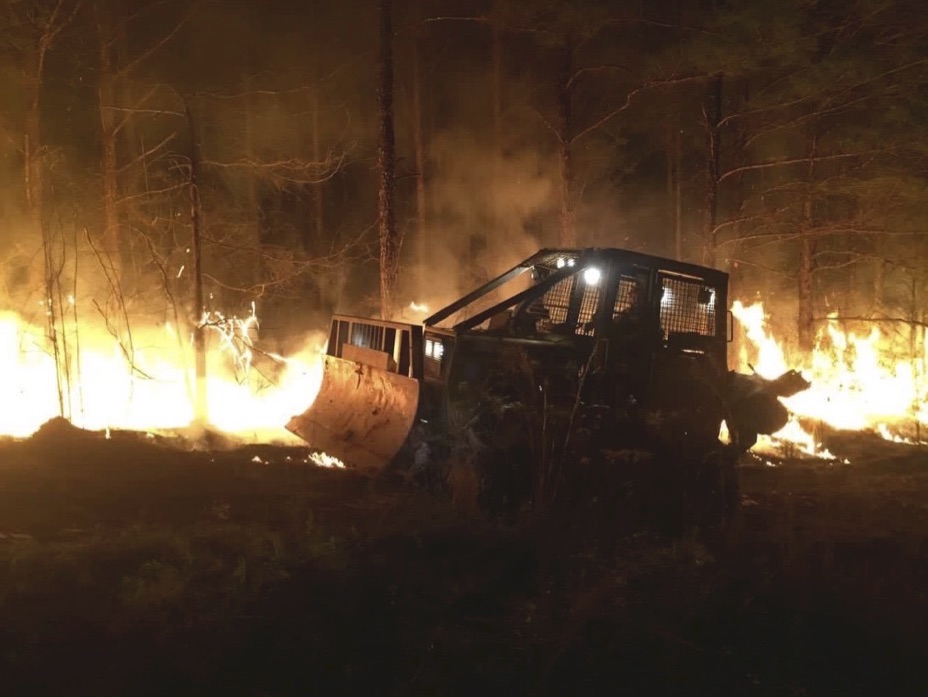 A small bulldozer driving at night, with a trees burning behind it.