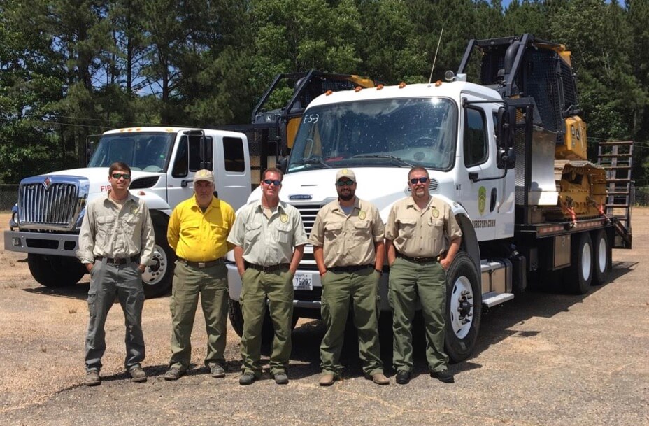 Five wildfire firefighters standing in front of two white trailer trucks with bulldozers loaded onto the trailer