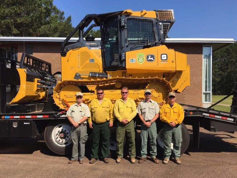 Five wildfire firefighters standing in front of a bright yellow bulldozer loaded onto a trailer bed as they prepare to deploy to Texas.