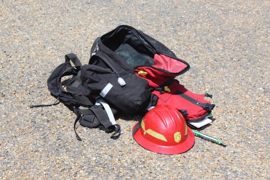 An open black backpack next to an open red bag on the ground. Sitting in front of them is a red fireman's hard hat