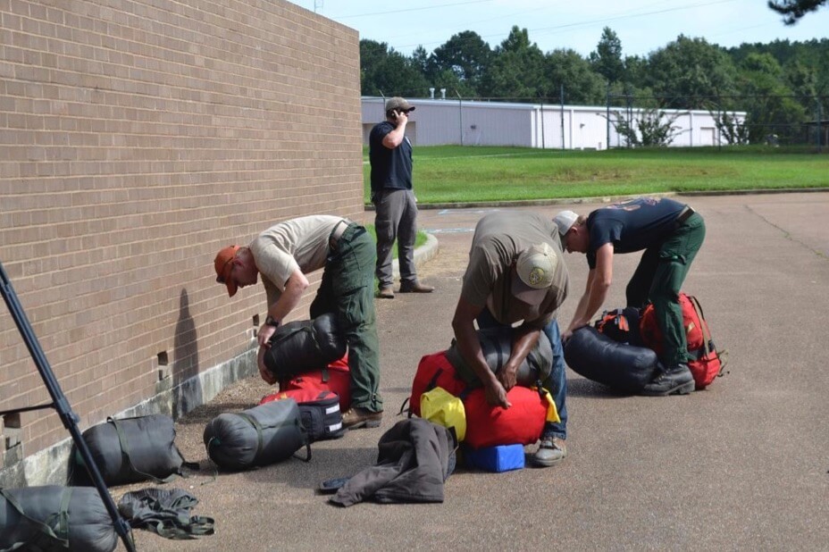 Three men packing backpacks while a man behind them talks on the phone