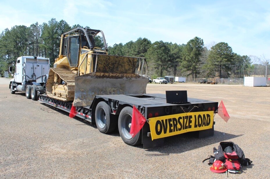 A trailer with the words "Oversize Load" and a bulldozer on it sitting behind an open firefighter's duffel bag.